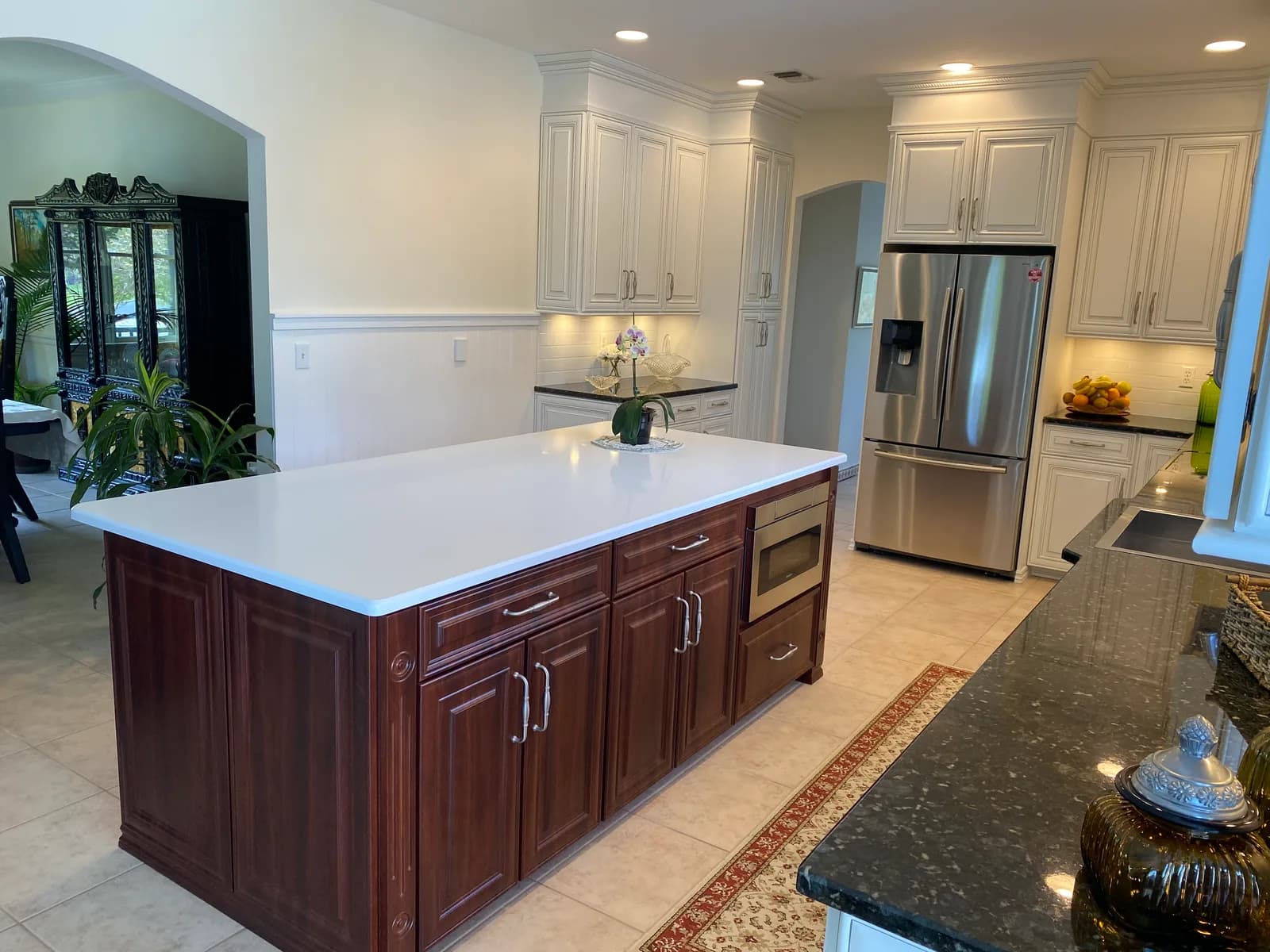 A kitchen with cream raised-panel perimeter cabinets and a dark cherry island topped with white quartz, beside a stainless refrigerator